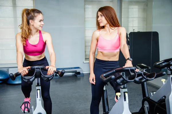 Dos hermosas mujeres, usando ropa deportiva, haciendo ejercicio de ciclismo en interiores . — Foto de Stock