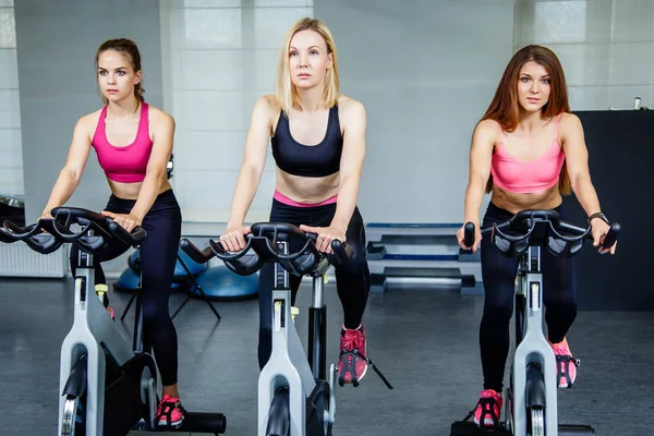 Tres hermosas mujeres jóvenes haciendo ejercicio en las bicicletas en el gimnasio — Foto de Stock