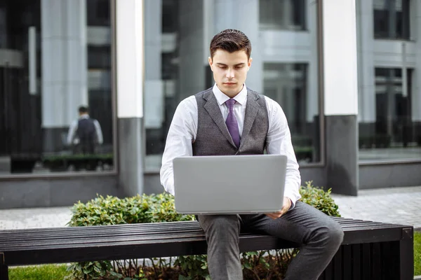 Молодой студент бизнес-школы работает за ноутбуком, сидя на скамейке — стоковое фото