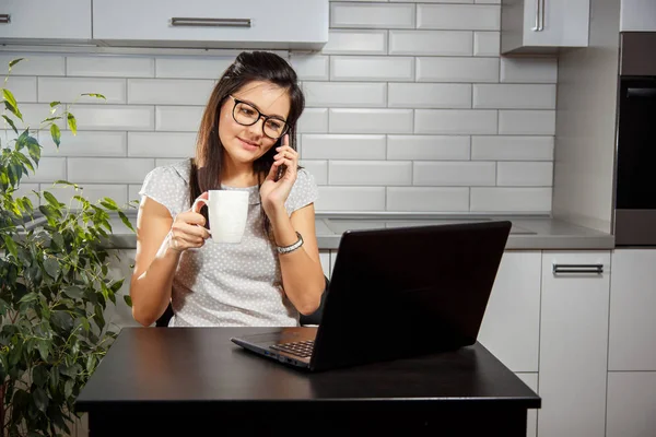 Молодая девушка разговаривает по телефону и пьет кофе сидя перед ноутбуком — стоковое фото