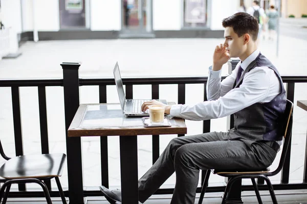 Молодой успешный бизнесмен сидит в открытом городском кафе с ноутбуком и разговаривает по телефону — стоковое фото