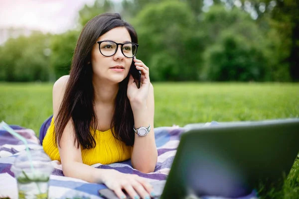 Çimenlerin üzerinde parlak yaz günü içinde oturan ve bilgisayarda çalışan gülümseyen kız. Dizüstü bilgisayar açık havada, telefonda konuşurken arkadaşlarınızla sohbet kullanarak. Sosyal ağ ve serbest.