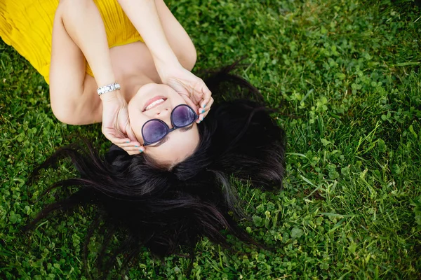 Happy young brunette woman in sunglasses lying on the grass, top view. — Stock Photo, Image