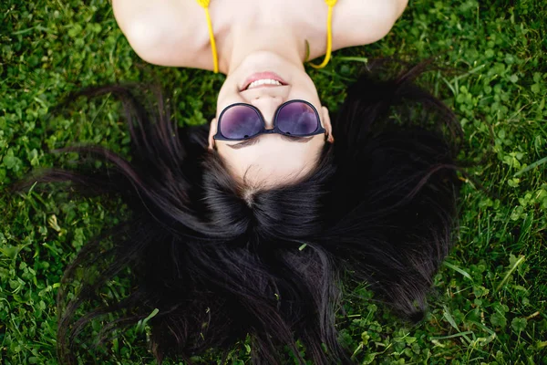 Happy young brunette woman in sunglasses lying on the grass, top view. — Stock Photo, Image