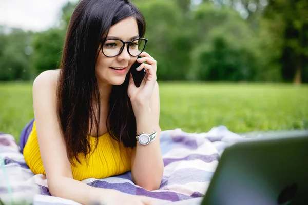 Chica sonriente sentada en el césped en un día brillante de verano y trabajando en la computadora. Usar el portátil al aire libre, chatear con amigos, hablar por teléfono. Redes sociales y freelance . — Foto de Stock