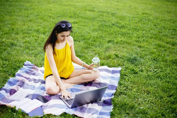 Красивая молодая женщина сидит на зеленой траве в парке со своим ноутбуком. Студентка смотрит в компьютер — стоковое фото
