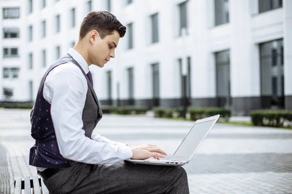 Молодой студент бизнес-школы, работающий за ноутбуком, сидя на скамейке запасных — стоковое фото