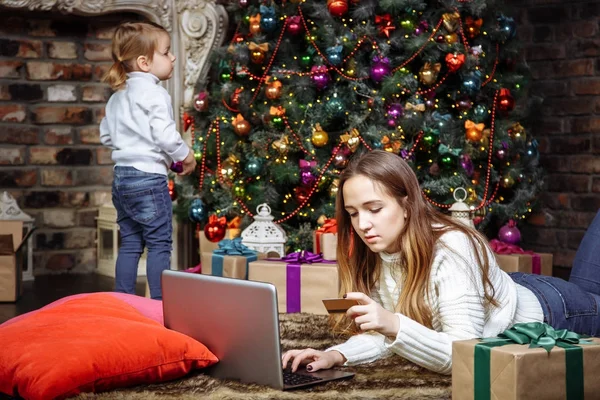 A young mother is doing online shopping with laptop and credit card while her daughter decorates the Christmas tree. The concept of preparing for Christmas and new year. — Stock Photo, Image