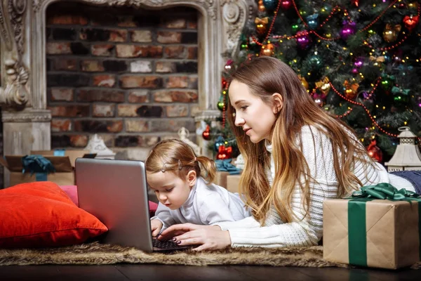A young mother and her daughter using a laptop lying near the Christmas tree — Stock Photo, Image