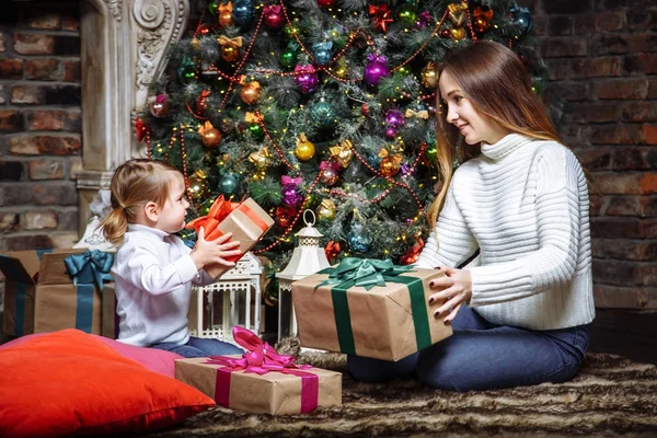 Merry Christmas and Happy Holidays! Cheerful mom and her cute daughter exchanging gifts. Parent and little child having fun near Christmas tree indoors. Loving family with presents in room. — Stock Photo, Image