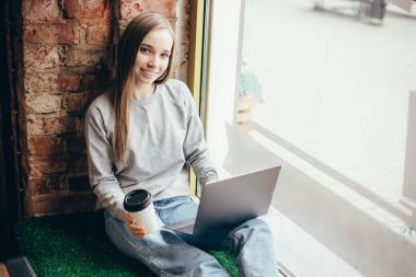 Pencere kenarında oturan ve dizüstü bilgisayar kullanarak güzel genç kadın