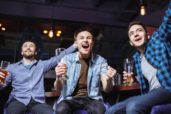 Three young men in casual clothes are cheering for football and holding glasses of beer while sitting at bar counter in pub