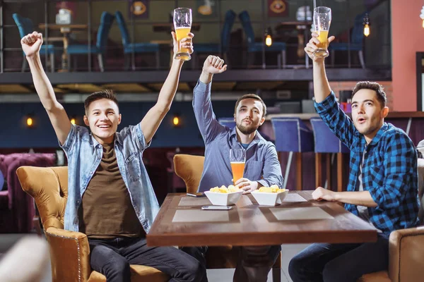 Trois jeunes hommes en vêtements décontractés applaudissent pour le football et tiennent des verres de bière tout en étant assis au bar . — Photo