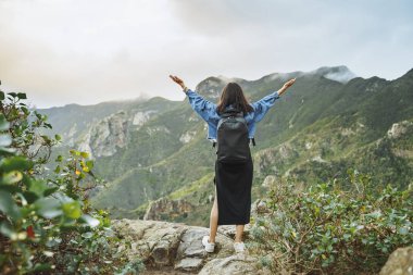 Sırt çantalı genç bir turist dağın tepesinde dinleniyor ve vadi manzarasının tadını çıkarıyor.
