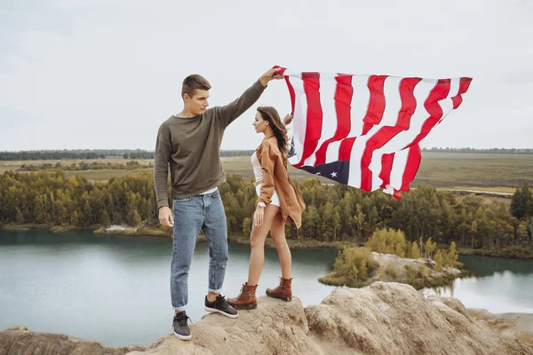 Felice giovane coppia che tiene una bandiera americana sventolando in natura. Giorno di indipendenza, stile di vita, concetto di viaggio — Foto Stock