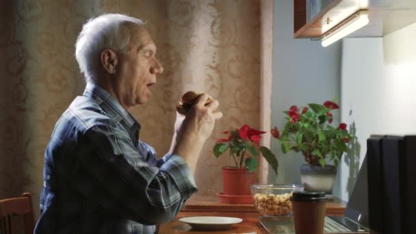 老年人晚上在家里用笔记本电脑看电影或电视节目 吃汉堡包 — 图库视频影像