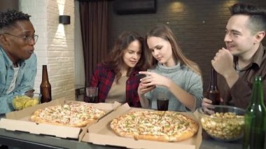 Mutlu çok ırklı arkadaşlar bira içiyor, pizza yiyor ve evdeki oturma odasında birlikte eğleniyorlar.