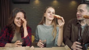 Bir grup arkadaş pizza yiyor, konuşuyor ve komik bir fıkraya gülüyorlar. Evdeki oturma odasında.