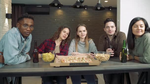 Група багатоетнічних друзів п'ють пиво і дивиться телевізор разом у вітальні вдома . — стокове відео