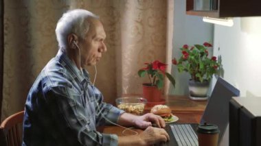 Kulaklıklı yaşlı bir adam bilgisayar kullanarak video bağlantısı kuruyor. Akşamları evdeki müdahalecisine başparmağını gösteriyor..