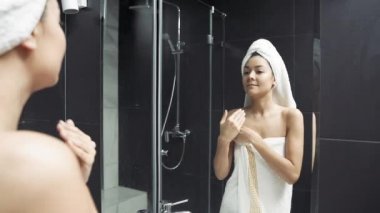 Kafasında havlu olan mutlu genç bir kadın banyoda vücut kremi sürüyor. Çekici esmer, bakımlı ve güzel teniyle aynaya bakıyor.