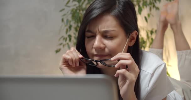若い女性は ベッドの上に寝そべってノートパソコンで作業しながら眼鏡を脱いで 長い着用後に不快感や眼の緊張を感じている 視力の問題 — ストック動画