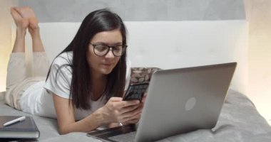 Evde çalışırken akıllı telefon ve dizüstü bilgisayar kullanarak yatakta yatan genç bir kadın.