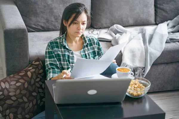 Young woman reading documents or reports while working on a laptop at home. Distance work or online education. Stay home while quarantined against coronavirus.