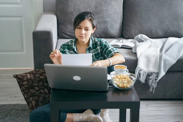 Young woman reading documents or reports while working on a laptop at home. Distance work or online education. Stay home while quarantined against coronavirus.