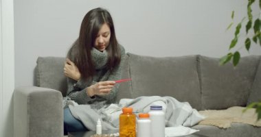 Genç hasta kadın termometreyi tutuyor, burnu akıyor ve evdeki kanepede oturuyor. Grip ya da virüs