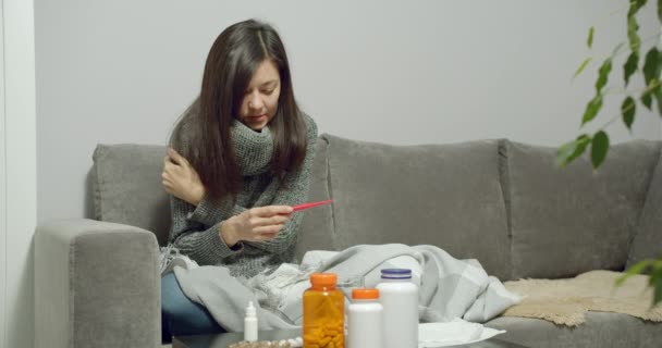 体温計を持っている若い病気の女性は自宅でソファに座って鼻水や寒気を実行しています インフルエンザやウイルス — ストック動画