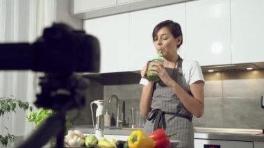 Çekici genç bir kadın yeni yapılmış smoothie 'yi tadıyor ve evdeki mutfakta dijital kamerada sağlıklı beslenmeyle ilgili bir video kaydediyor. Vlog ve sosyal medya kavramı.