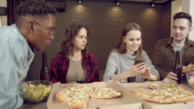 Bir grup çok ırklı arkadaş pizza yiyor, bira içiyor ve evdeki oturma odasında sohbet ediyorlar..