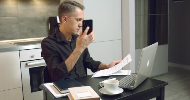 若い男は契約書やレポートを読んで電話で話しています 家のオフィスからノートパソコンで働くビジネスマン — ストック動画