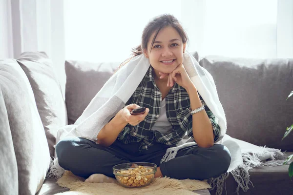 若いです笑顔女性ラップでプレイした食べるポップコーンと見てテレビながら座っている間にソファに家 ロイヤリティフリーのストック画像