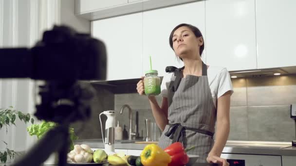 Vonzó fiatal nő kóstolja meg a frissen készített smoothie-t, és rögzít egy videót az egészséges étkezésről digitális fényképezőgépen otthon a konyhában. Vlogging és a közösségi média koncepciója.