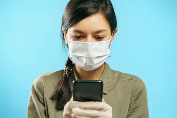 青い背景にスマートフォンを使って保護医療用マスクや手袋をした若い女性 オンラインチャット ロイヤリティフリーのストック画像