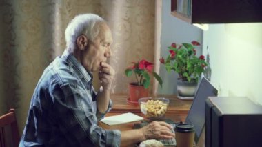 Yaşlı bir adam akşam evde dizüstü bilgisayarla çalışırken patlamış mısır yiyor ve klavyede daktilo kullanıyor. Çevrimiçi iletişim ya da uzak çalışma