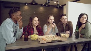 Bir grup mutlu çoklu etnik arkadaş pizza yiyor, bira içiyor ve evdeki oturma odasında televizyon seyrediyor.