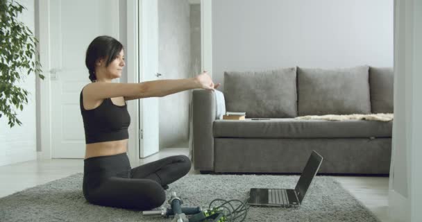 自宅でラップトップでフィットネスビデオをオンラインで見ている間にストレッチ演習を行うスポーティ若い女性 健康的なライフスタイルのコンセプト 自己分離中のスポーツ — ストック動画