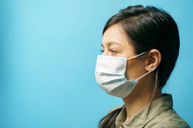 Mavi arka planda koruyucu maske takan üzgün Asyalı genç bir kadının portresi. Korku ve yalnızlık kavramı. Boşluğu kopyala