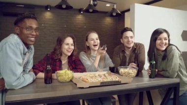 Bir grup mutlu çoklu-etnik arkadaş pizza yiyip bira içerek komik bir şakaya gülüyorlar ve evdeki oturma odasında birlikte televizyon izliyorlar.
