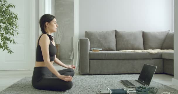 自宅のラップトップでフィットネスビデオをオンラインで見ている間 蓮の位置に座って深く呼吸するスポーティ若い女性 健康的なライフスタイルのコンセプト 自己分離中のリラクゼーション — ストック動画