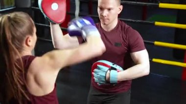 Boks eldivenli genç bir kadın boks hocasını yumrukluyor. Boks tekmelerini koruma amaçlı kullanıyor..