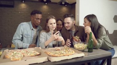 Beyaz bir kadın, arkadaşlarıyla birlikte bira içip pizza yerken ya da barda bir şeyler yerken akıllı telefonuyla arkadaşlarına bir şey gösteriyor..
