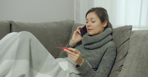 体温を測定し 自宅でソファに座っている間に相談のために医師を呼び出す病気の若い女性の肖像画 — ストック動画