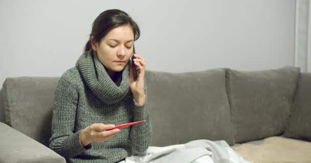 体温を測定し 自宅でソファに座っている間に相談のために医師を呼び出す病気の若い女性の肖像画 — ストック動画