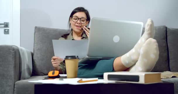 同僚やクライアントのために音声メッセージを録音した眼鏡の若い女性自宅のソファに座ってノートパソコンで作業する紙の上に計画や図面を分析する — ストック動画
