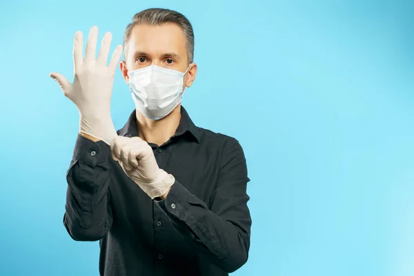 Portrait of a young man in a medical mask putting on protective gloves on a blue background.