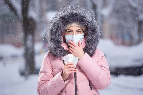 Retrato de uma menina bonita em uma jaqueta rosa em uma máscara protetora médica em segurar branco, rosa, pílulas amarelas em suas mãos para resfriados e gripe. Retrato de rua de inverno de uma mulher sob queda de neve — Fotografia de Stock